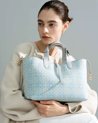 Blue floral brocade tote bag with leather handles and gold hardware, front half-body view of female model in beige outfit