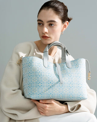 Blue floral brocade tote bag with leather handles and gold hardware, front half-body view of female model in beige outfit