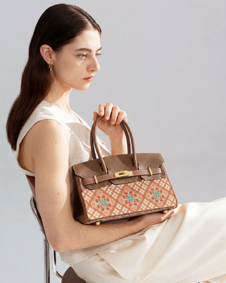 Brown caisson-pattern brocade & leather handbag with gold hardware, side half-body view of female model in white outfit