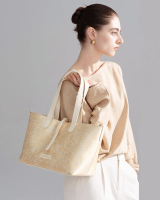 Gold brocade tote bag with leather handles, side half-body view of female model in beige top and white trousers