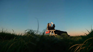 Side full-body view of female model in black dress, seated outdoors at sunset with a red embroidered hobo bag resting on the chair