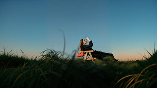 Side full-body view of female model in black dress, seated outdoors at sunset with a red embroidered hobo bag resting on the chair