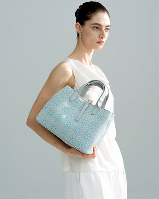 Side half-body view of model in white outfit carrying the silkyhaus light blue patterned silk brocade & genuine leather tote bag with gold hardware